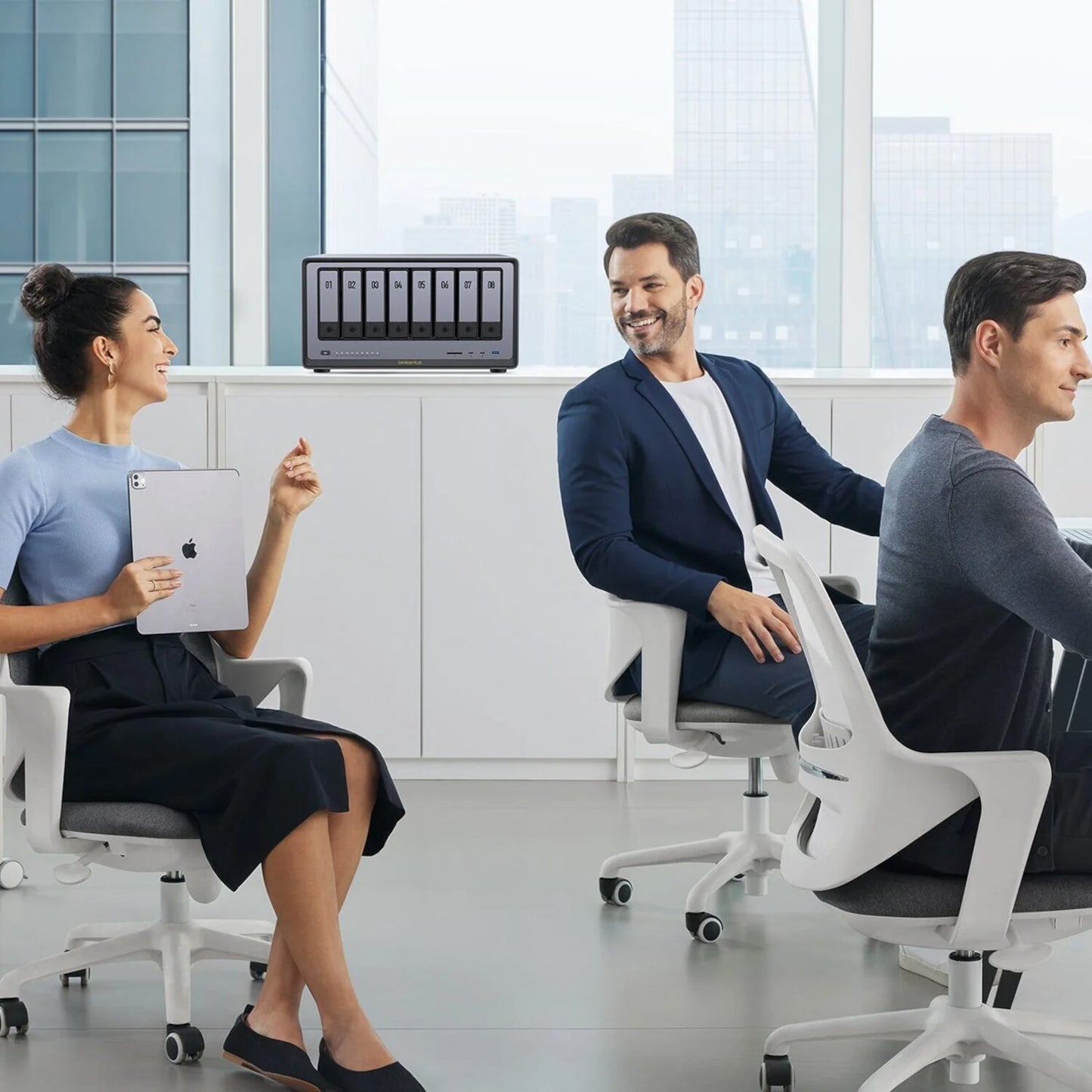 Three people in a modern office setting with a woman holding a tablet and two men sitting on chairs.