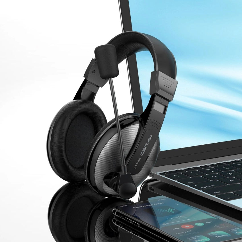 Black gaming headset on a reflective surface with a blurred laptop and phone in the background