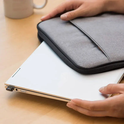 A gray fabric laptop sleeve with black trim holding a white laptop on a light wood desk, with a person's hands holding the sleeve and laptop.