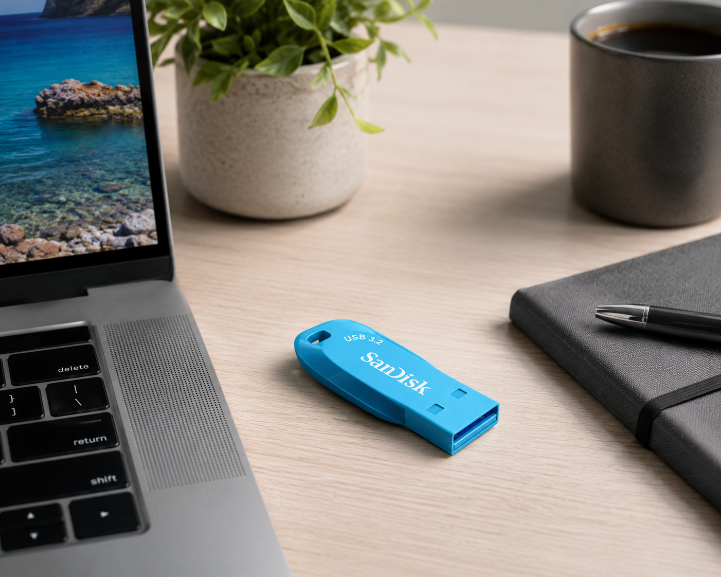 A bright blue SanDisk USB flash drive on a light wood desk next to a laptop, notebook, pen, and a cup of coffee, with a potted plant in the background.