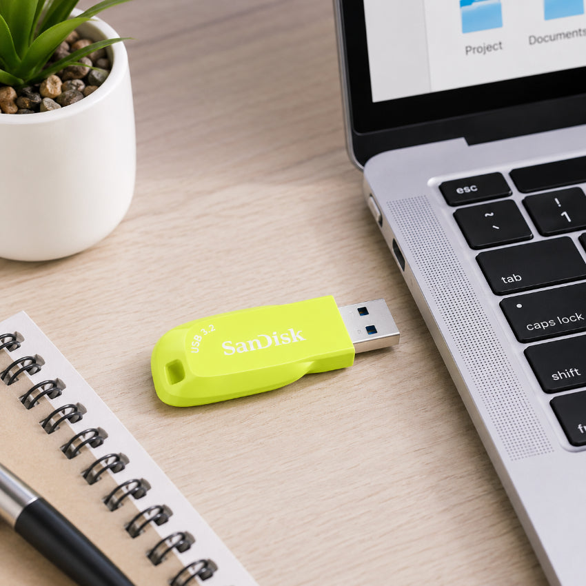 A bright yellow SanDisk Ultra Shift USB flash drive lies on a light wood desk next to a laptop, notebook, pen, and a small potted succulent.