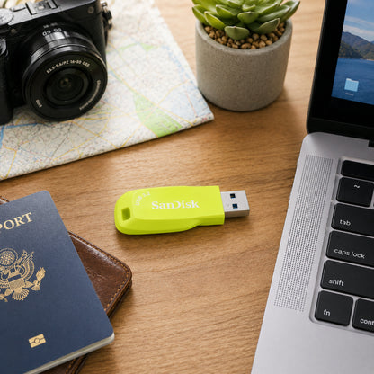 A bright yellow SanDisk USB flash drive lies on a wooden desk surrounded by everyday items including a passport, camera, laptop, and small potted succulent.
