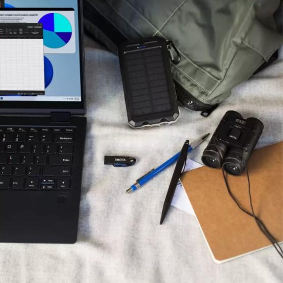 A top-down flat lay of outdoor and tech accessories on a light fabric surface, including a black laptop, a rugged solar power bank, a green backpack, black binoculars, a USB flash drive, a blue pen, a black pen, and a brown notebook.
