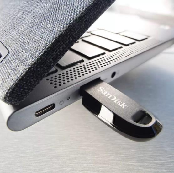 A close-up of a SanDisk USB flash drive inserted into a silver laptop, with a dark textured fabric sleeve partially visible on the left.
