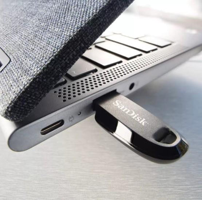 A close-up of a SanDisk USB flash drive inserted into a silver laptop, with a dark textured fabric sleeve partially visible on the left.