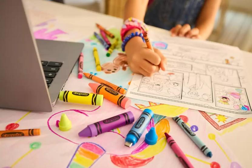 A child's hand drawing on a coloring page at a table covered with crayons, a laptop, and colorful artwork, with a person partially visible in the background.