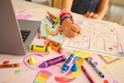 A child's hand drawing on a coloring page at a table covered with crayons, a laptop, and colorful artwork, with a person partially visible in the background.