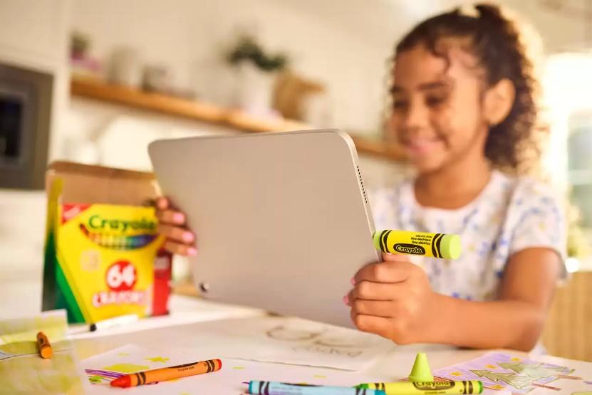 A young girl with curly hair smiles while holding a tablet and a yellow Crayola crayon at a table with drawing papers and other Crayola crayons in a brightly lit room.
