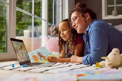 A woman and a young girl are lying on a bed looking at a laptop screen while reading a colorful children's book. The room has a large window with daylight, and toys and drawings are scattered on the bed.