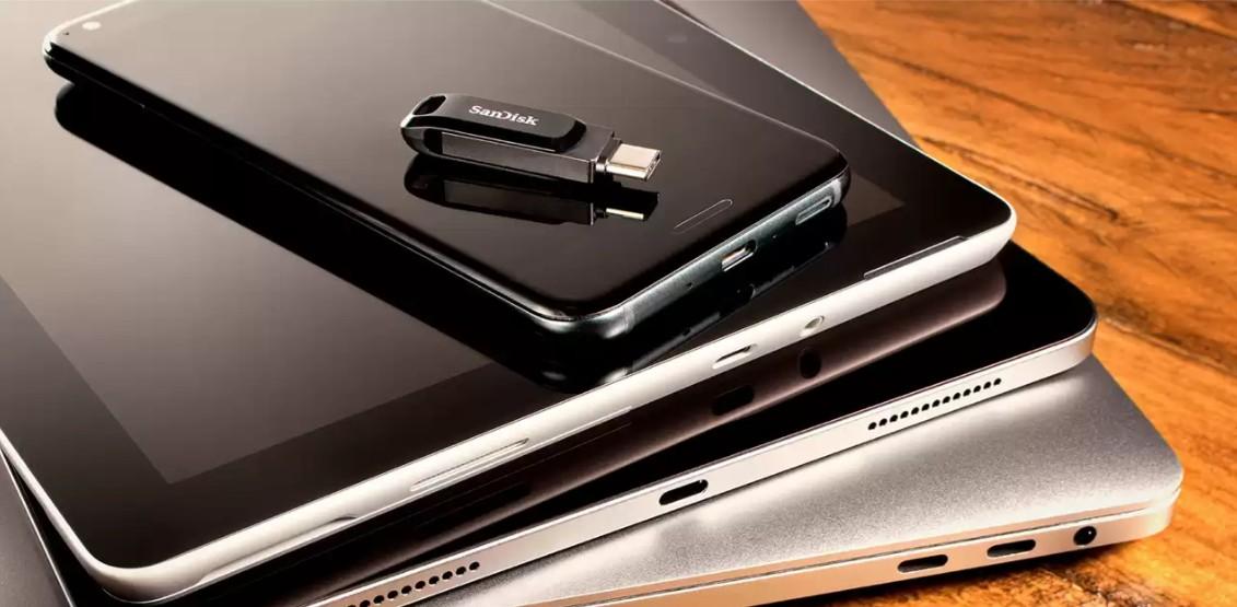 A SanDisk USB flash drive is shown resting on the back of a black tablet, with two silver tablets stacked underneath on a wooden surface.