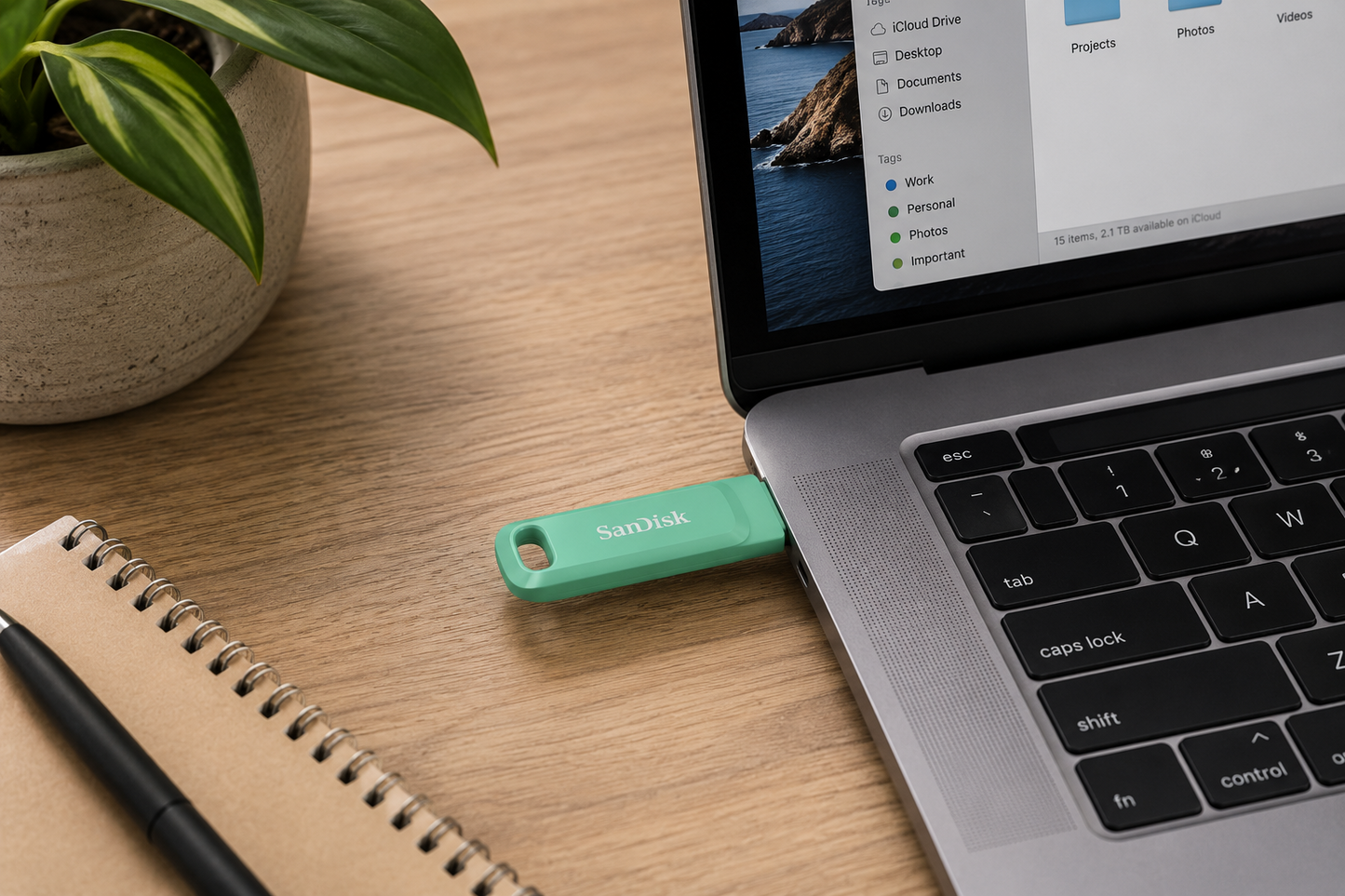 A mint green SanDisk USB flash drive plugged into a silver laptop on a wooden desk, with a notebook, pen, and potted plant nearby.