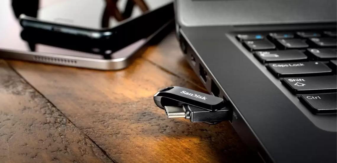 A SanDisk USB flash drive plugged into a black laptop on a wooden desk, with a smartphone blurred in the background.