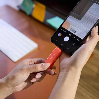 Person holding a Sandisk USB Flash Drive next to a smartphone on a desk.