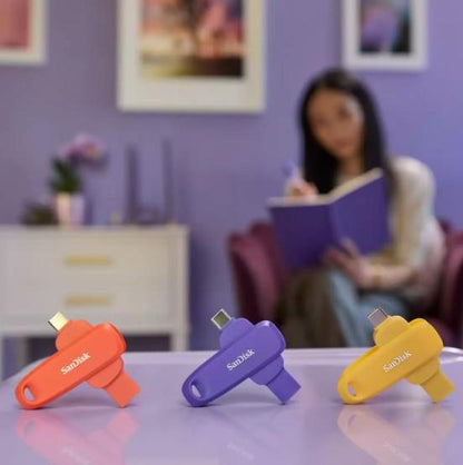Three SanDisk USB flash drives in orange, purple, and yellow are displayed on a reflective purple table, with a blurred woman reading in a room with a purple wall in the background.
